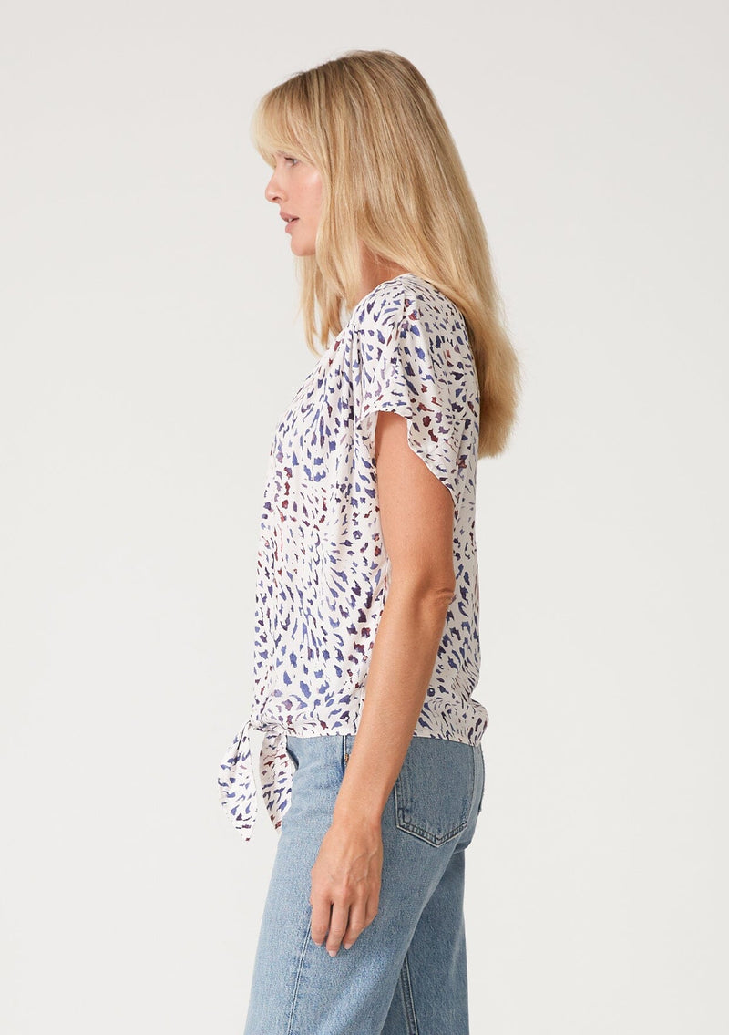 [Color: Natural/Ink] A side facing image of a blonde model wearing a casual resort top in a multi colored abstract print. With short sleeves, a loop button front, a v neckline, and a tie front waist detail. 