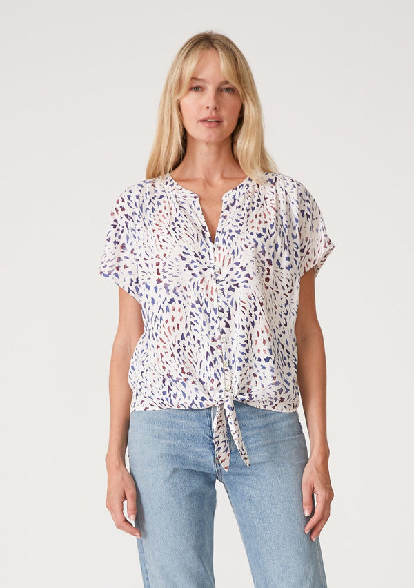 [Color: Natural/Ink] A front facing image of a blonde model wearing a casual resort top in a multi colored abstract print. With short sleeves, a loop button front, a v neckline, and a tie front waist detail. 