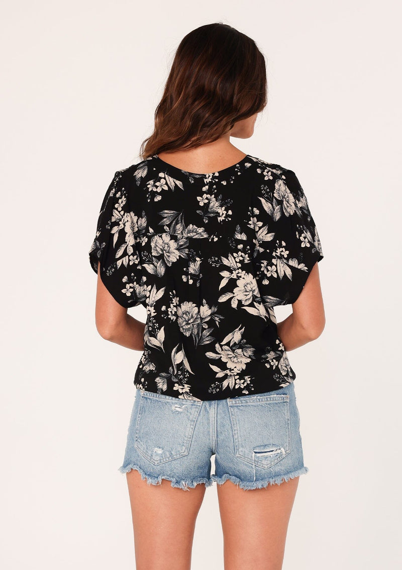 [Color: Black/Natural] A back facing image of a brunette model wearing a casual short sleeve top in a black and off white floral print. With a loop button front, a v neckline, and a tie front waist. 