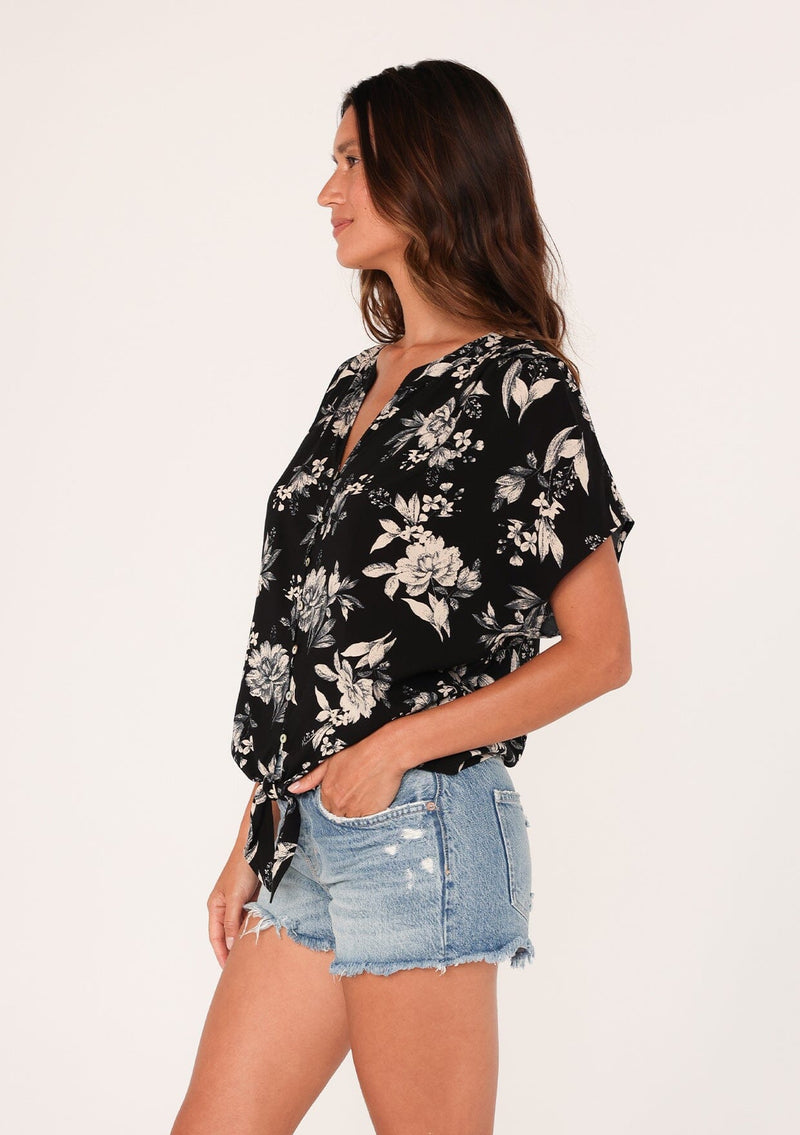 [Color: Black/Natural] A side facing image of a brunette model wearing a casual short sleeve top in a black and off white floral print. With a loop button front, a v neckline, and a tie front waist. 