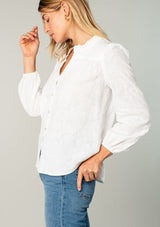 [Color: White] A side facing image of a blonde model wearing a classic bohemian cotton white button front shirt in a textured jacquard. With long sleeves and a ruffled neckline.