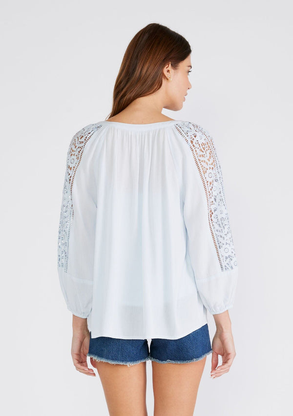 [Color: Ice Blue] A back facing image of a brunette model wearing a light blue bohemian resort blouse with voluminous long sleeves, a button front, tassel neck ties, and lace trim throughout.