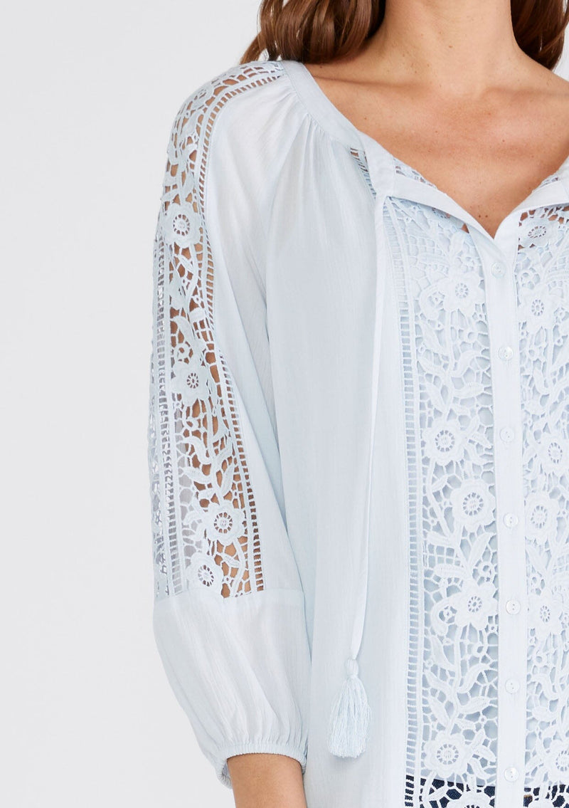 [Color: Ice Blue] A close up front facing image of a brunette model wearing a light blue bohemian resort blouse with voluminous long sleeves, a button front, tassel neck ties, and lace trim throughout.