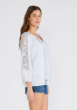 [Color: Ice Blue] A side facing image of a brunette model wearing a light blue bohemian resort blouse with voluminous long sleeves, a button front, tassel neck ties, and lace trim throughout.