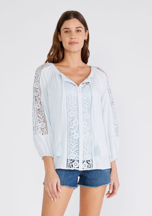 [Color: Ice Blue] A front facing image of a brunette model wearing a light blue bohemian resort blouse with voluminous long sleeves, a button front, tassel neck ties, and lace trim throughout.