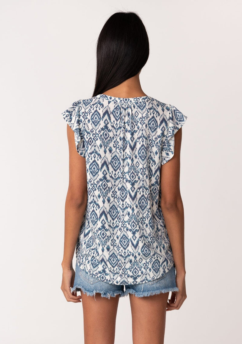 [Color: Ivory/Denim] A back facing image of a brunette model wearing a casual bohemian blouse in a blue bohemian diamond print. With short ruffle sleeves, a v neckline, and a relaxed fit. 