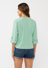 [Color: Seafoam Green] A back facing image of a brunette model wearing a green cotton jacquard shirt with a split v-neckline, concealed button front, tie front detail, and long rolled sleeves with button tab. 