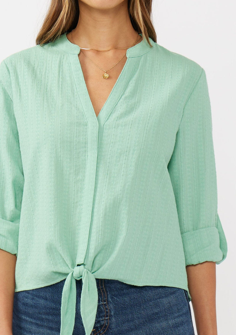 [Color: Seafoam Green] A detail image of a brunette model wearing a green cotton jacquard shirt with a split v-neckline, concealed button front, tie front detail, and long rolled sleeves with button tab. 