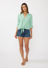 [Color: Seafoam Green] A front facing image of a brunette model wearing a green cotton jacquard shirt with a split v-neckline, concealed button front, tie front detail, and long rolled sleeves with button tab. 