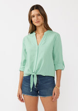 [Color: Seafoam Green] A front facing image of a brunette model wearing a green cotton jacquard shirt with a split v-neckline, concealed button front, tie front detail, and long rolled sleeves with button tab. 