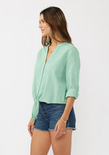 [Color: Seafoam Green] A side facing image of a brunette model wearing a green cotton jacquard shirt with a split v-neckline, concealed button front, tie front detail, and long rolled sleeves with button tab. 