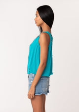 [Color: Turquoise] A side facing image of a brunette model wearing a casual turquoise blue bohemian summer tank top with a surplice v neckline, an elastic waist, and a back soutache braided detail. 