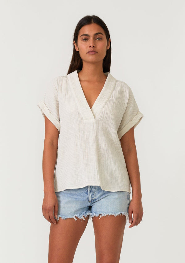 [Color: Cream] A front facing image of a brunette model wearing an off white cotton gauze tee. With short cuffed sleeves, a v neckline, and an oversized relaxed fit. 