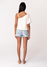 [Color: White/Apricot] A back facing image of a brunette model wearing a lightweight cotton summer blouse in a white and pink embroidered design. With a one shoulder asymmetric neckline, a short puff sleeve, an adjustable spaghetti strap, and a relaxed fit. 