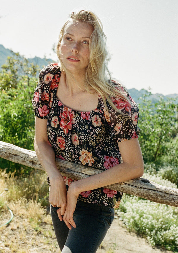 [Color: Black/Fuchsia] A half body front facing image of a blonde model standing outside wearing a bohemian spring top in a black and pink floral print. With short puff sleeves, a square neckline, and a relaxed fit.