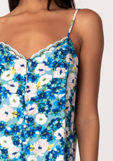 [Color: Dusty Teal/Blue] A close up front facing image of a brunette model wearing a realxed fit sleeveless camisole tank top in a blue floral print. With adjustable spaghetti straps, a loop button front, a scallop trimmed hemline, and a scallop trimmed v neckline. 