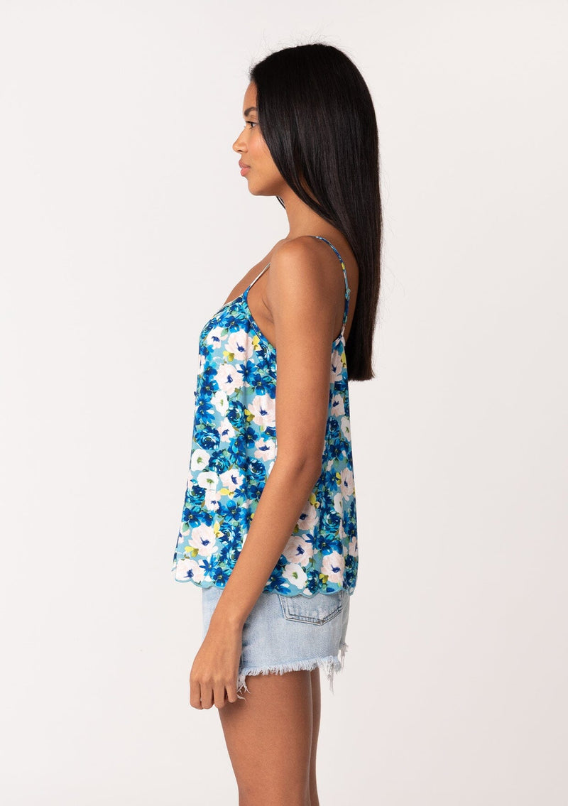 [Color: Dusty Teal/Blue] A side facing image of a brunette model wearing a realxed fit sleeveless camisole tank top in a blue floral print. With adjustable spaghetti straps, a loop button front, a scallop trimmed hemline, and a scallop trimmed v neckline. 