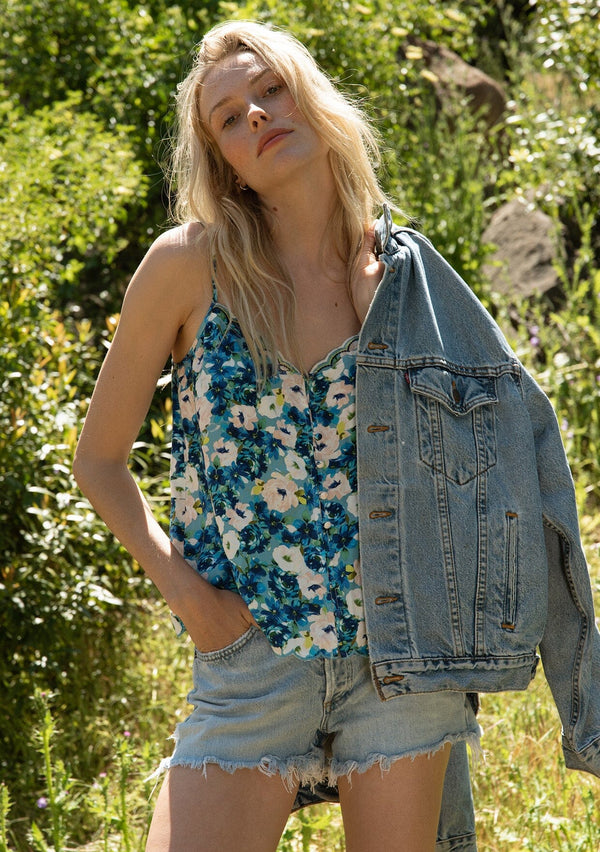 [Color: Dusty Teal/Blue] A half body front facing image of a blonde model standing in a field wearing a realxed fit sleeveless camisole tank top in a blue floral print. With adjustable spaghetti straps, a loop button front, a scallop trimmed hemline, and a scallop trimmed v neckline.