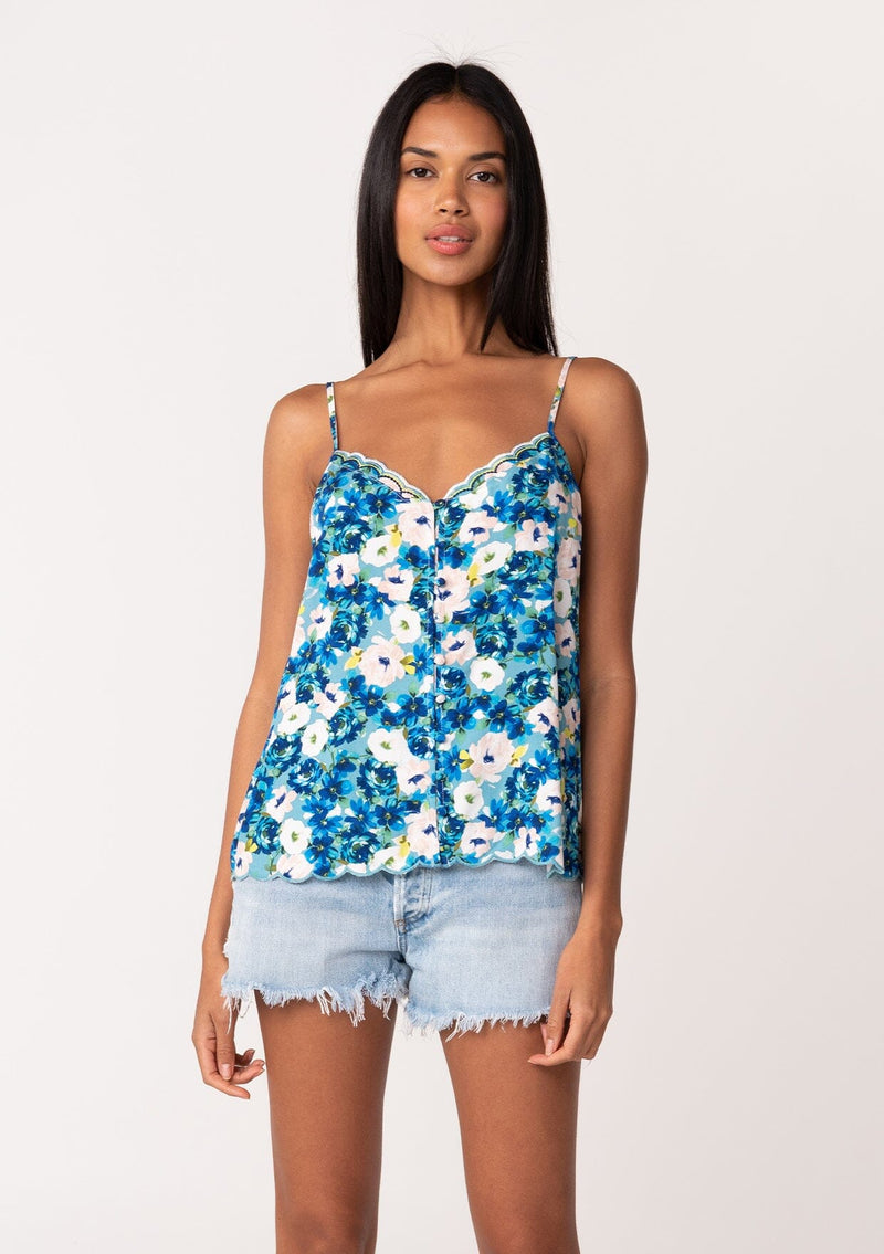 [Color: Dusty Teal/Blue] A front facing image of a brunette model wearing a realxed fit sleeveless camisole tank top in a blue floral print. With adjustable spaghetti straps, a loop button front, a scallop trimmed hemline, and a scallop trimmed v neckline. 
