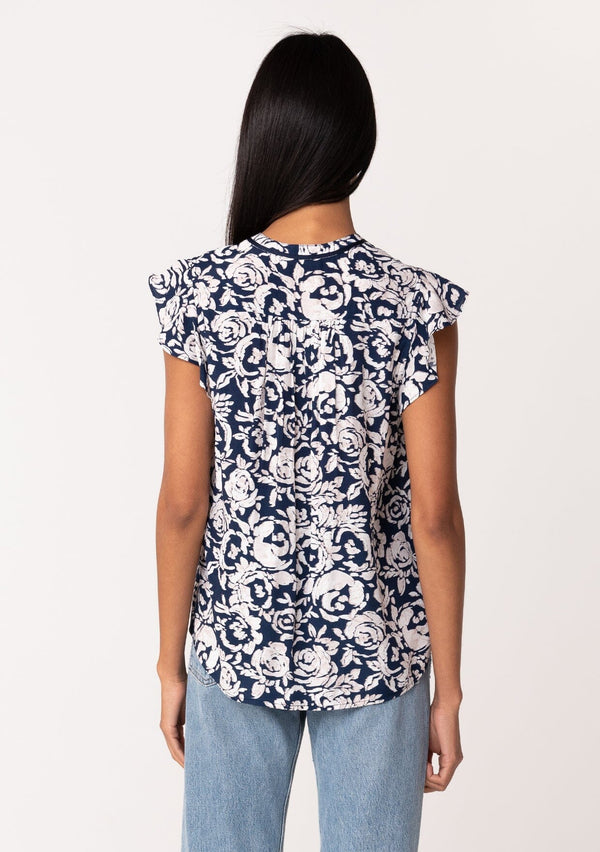 [Color: Navy/Natural] A back facing image of a brunette model wearing a blue floral summer blouse. With short flutter sleeves, a split v neckline with double tassel ties, and a relaxed fit. 