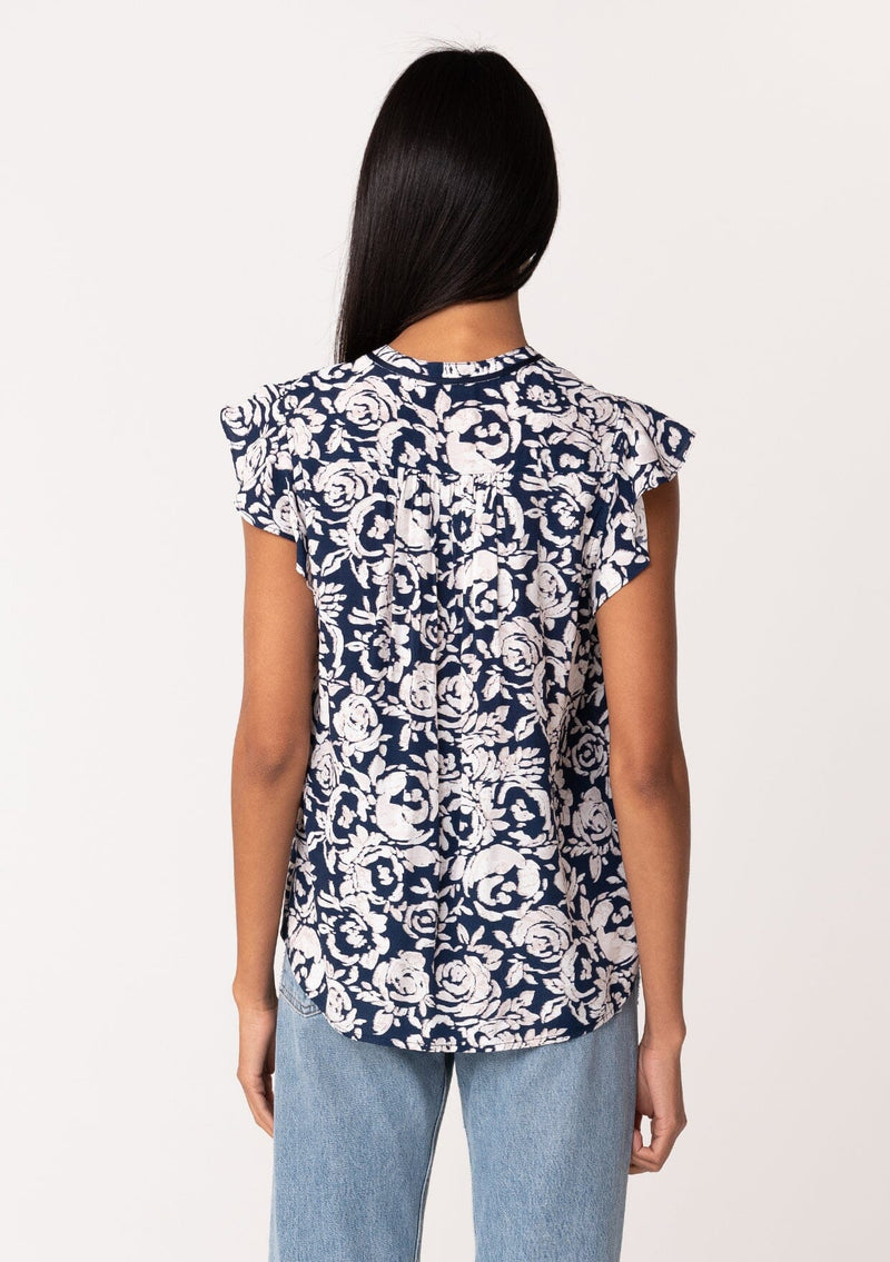 [Color: Navy/Natural] A back facing image of a brunette model wearing a blue floral summer blouse. With short flutter sleeves, a split v neckline with double tassel ties, and a relaxed fit. 