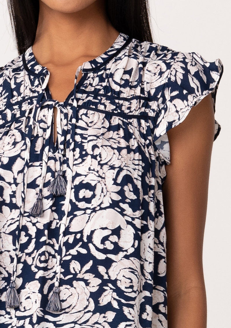 [Color: Navy/Natural] A close up front facing image of a brunette model wearing a blue floral summer blouse. With short flutter sleeves, a split v neckline with double tassel ties, and a relaxed fit. 