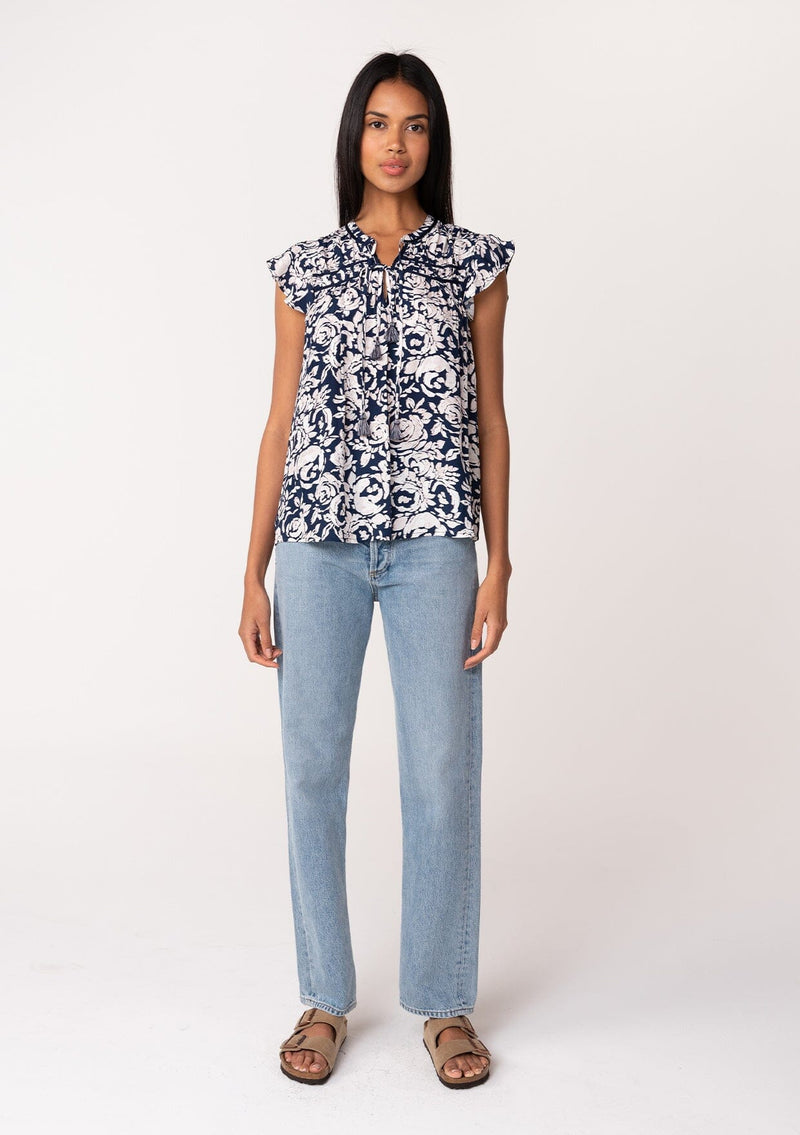 [Color: Navy/Natural] A full body front facing image of a brunette model wearing a blue floral summer blouse. With short flutter sleeves, a split v neckline with double tassel ties, and a relaxed fit. 