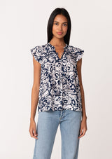 [Color: Navy/Natural] A front facing image of a brunette model wearing a blue floral summer blouse. With short flutter sleeves, a split v neckline with double tassel ties, and a relaxed fit. 