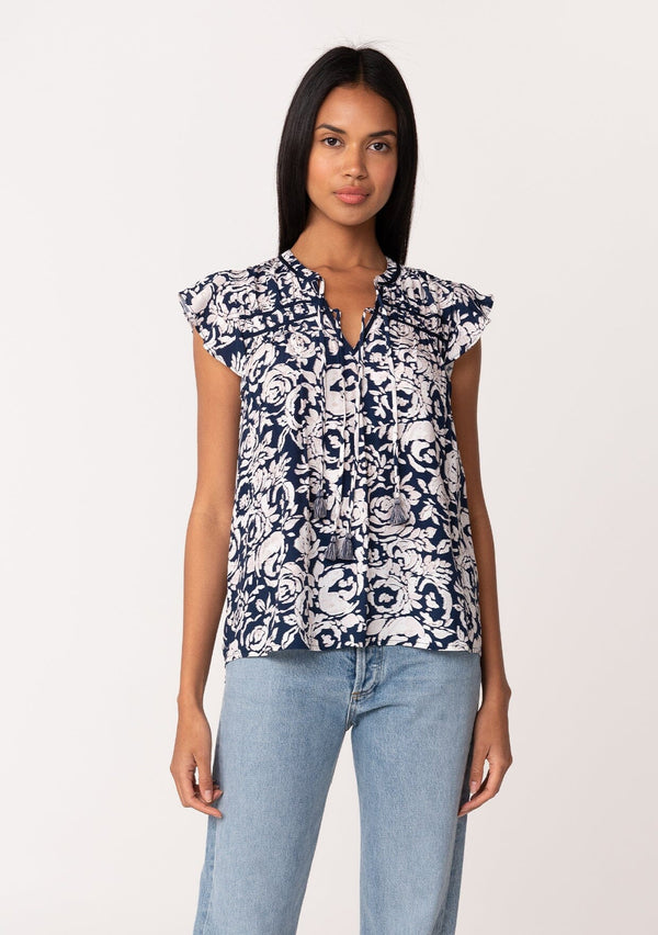 [Color: Navy/Natural] A front facing image of a brunette model wearing a blue floral summer blouse. With short flutter sleeves, a split v neckline with double tassel ties, and a relaxed fit. 