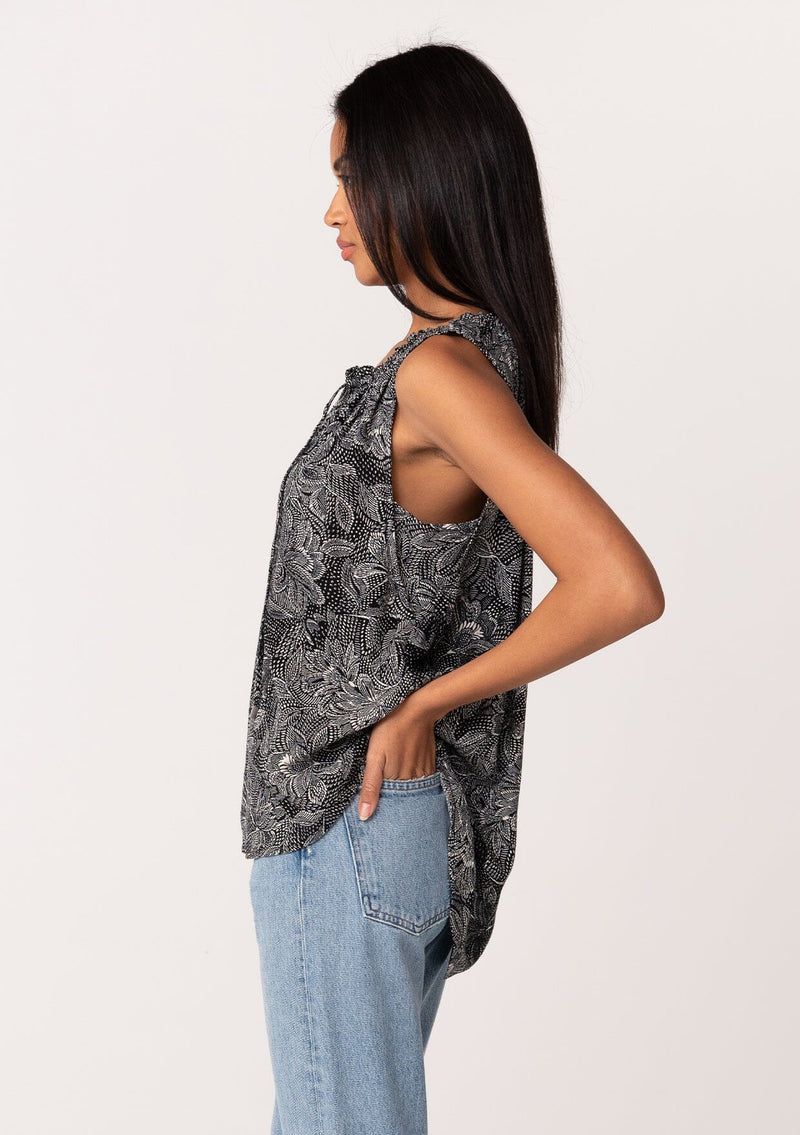 [Color: Black/Natural] A side facing image of a brunette model wearing a lightweight sleeveless blouse in a black and natural floral print. With a ruffled neckline, a split v-neckline with ties, and a relaxed fit. 