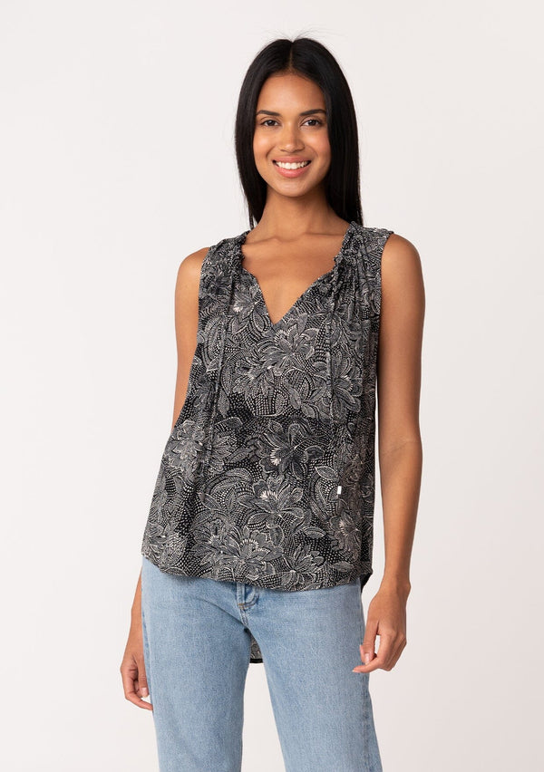 [Color: Black/Natural] A front facing image of a brunette model wearing a lightweight sleeveless blouse in a black and natural floral print. With a ruffled neckline, a split v-neckline with ties, and a relaxed fit. 