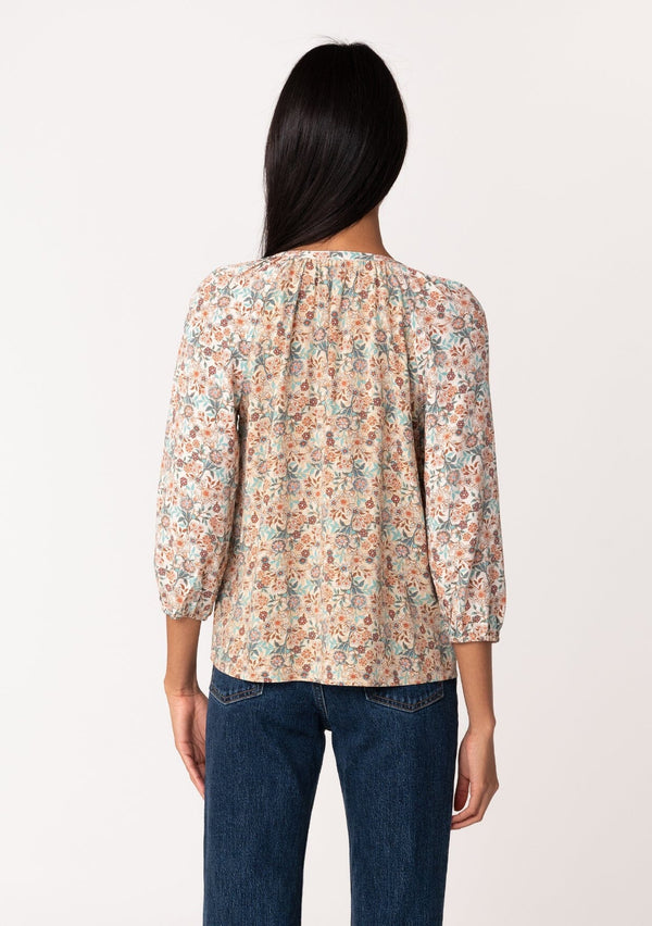 [Color: Natural/Teal] A back facing image of a brunette model wearing a fall blouse in a natural floral print. With three quarter length sleeves, a self covered button front, a split v neckline with ties, and a relaxed fit. 
