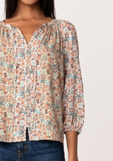 [Color: Natural/Teal] A close up front facing image of a brunette model wearing a fall blouse in a natural floral print. With three quarter length sleeves, a self covered button front, a split v neckline with ties, and a relaxed fit. 