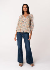 [Color: Natural/Teal] A full body front facing image of a brunette model wearing a fall blouse in a natural floral print. With three quarter length sleeves, a self covered button front, a split v neckline with ties, and a relaxed fit. 
