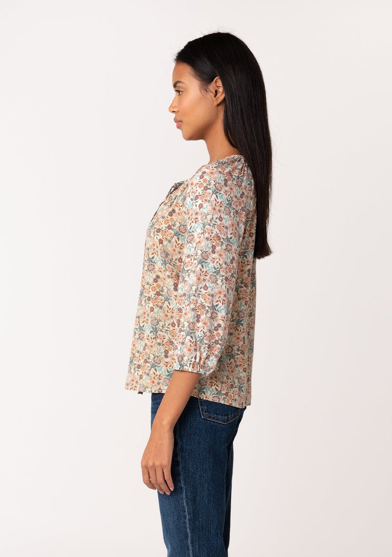 [Color: Natural/Teal] A side facing image of a brunette model wearing a fall blouse in a natural floral print. With three quarter length sleeves, a self covered button front, a split v neckline with ties, and a relaxed fit. 