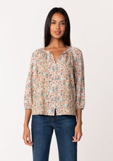 [Color: Natural/Teal] A front facing image of a brunette model wearing a fall blouse in a natural floral print. With three quarter length sleeves, a self covered button front, a split v neckline with ties, and a relaxed fit. 
