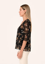 [Color: Black/Peach] A side facing image of a blonde model wearing a delicate chiffon bohemian blouse in a black and peach floral print. With half length flutter sleeves, a v neckline, and a self covered loop button front. 