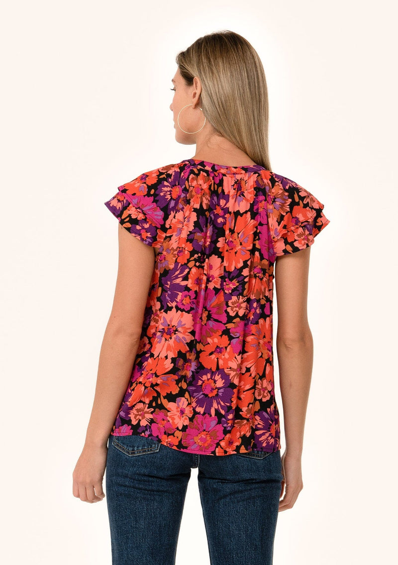 [Color: Black/Fuchsia] A back facing image of a blonde model wearing a fall bohemian blouse in a bright pink floral print. With a double flutter cap sleeve, a v neckline with ties, and a self covered loop button front. 