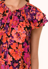 [Color: Black/Fuchsia] A close up front facing image of a blonde model wearing a fall bohemian blouse in a bright pink floral print. With a double flutter cap sleeve, a v neckline with ties, and a self covered loop button front. 