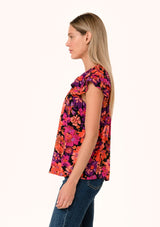 [Color: Black/Fuchsia] A side facing image of a blonde model wearing a fall bohemian blouse in a bright pink floral print. With a double flutter cap sleeve, a v neckline with ties, and a self covered loop button front. 