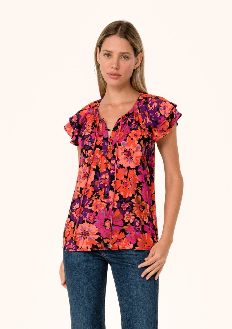 [Color: Black/Fuchsia] A front facing image of a blonde model wearing a fall bohemian blouse in a bright pink floral print. With a double flutter cap sleeve, a v neckline with ties, and a self covered loop button front. 