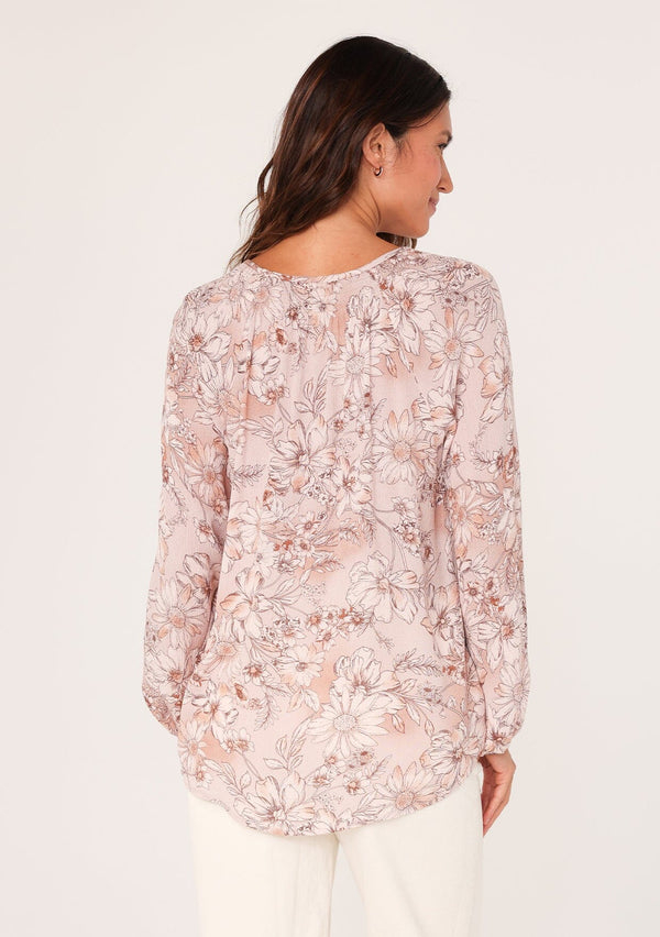 [Color: Dusty Rose/Light Brown] A back facing image of a brunette model wearing a boho blouse in a pink floral print. With long sleeves, a self covered button front, a split v neckline with tassel ties, and smocked details along the neckline. 