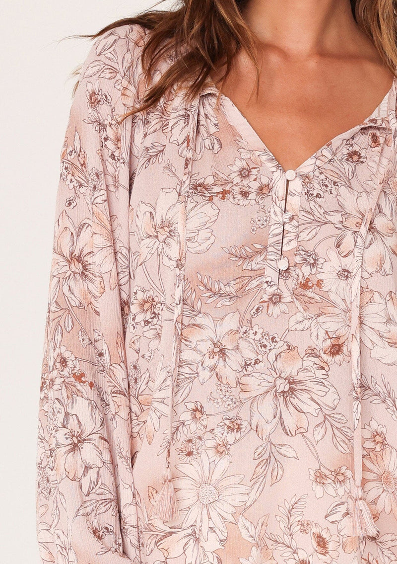 [Color: Dusty Rose/Light Brown] A close up front facing image of a brunette model wearing a boho blouse in a pink floral print. With long sleeves, a self covered button front, a split v neckline with tassel ties, and smocked details along the neckline. 