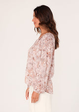 [Color: Dusty Rose/Light Brown] A side facing image of a brunette model wearing a boho blouse in a pink floral print. With long sleeves, a self covered button front, a split v neckline with tassel ties, and smocked details along the neckline. 