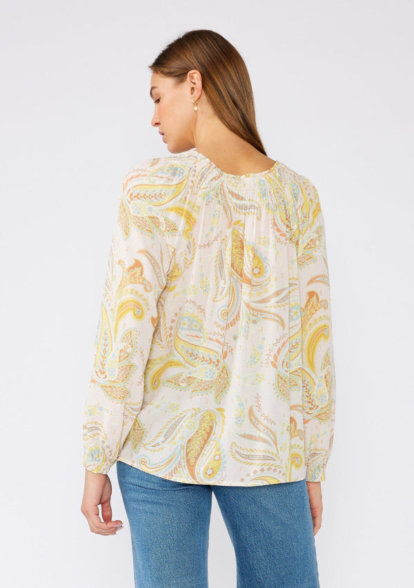 [Color: Dusty Peach/Caramel] A back facing image of a brunette model wearing a bohemian blouse in a pink and yellow paisley print. With gold lurex details, long raglan sleeves, a split v neckline with optional hook and eye closure, neck ties, and smocked details at the neckline. 