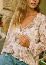 [Color: Natural/Dusty Rose] A close up front facing image of a blonde model wearing a fall bohemian blouse in an ivory and dusty pink paisley print. With gold metallic details throughout, three quarter length raglan sleeves, ruffled elastic wrist cuffs, a smocked neck, and a v neckline with ties.