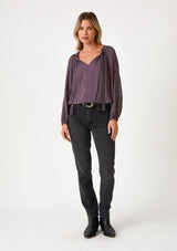 [Color: Dusty Plum] A full body front facing image of a blonde model wearing a dusty purple bohemian blouse with a sparkly sequined stripe. With long raglan sleeves, a split v neckline with tassel ties, and a ruffled elastic hemline. 