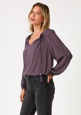 [Color: Dusty Plum] A front facing image of a blonde model wearing a dusty purple bohemian blouse with a sparkly sequined stripe. With long raglan sleeves, a split v neckline with tassel ties, and a ruffled elastic hemline. 