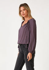 [Color: Dusty Plum] A side facing image of a blonde model wearing a dusty purple bohemian blouse with a sparkly sequined stripe. With long raglan sleeves, a split v neckline with tassel ties, and a ruffled elastic hemline. 