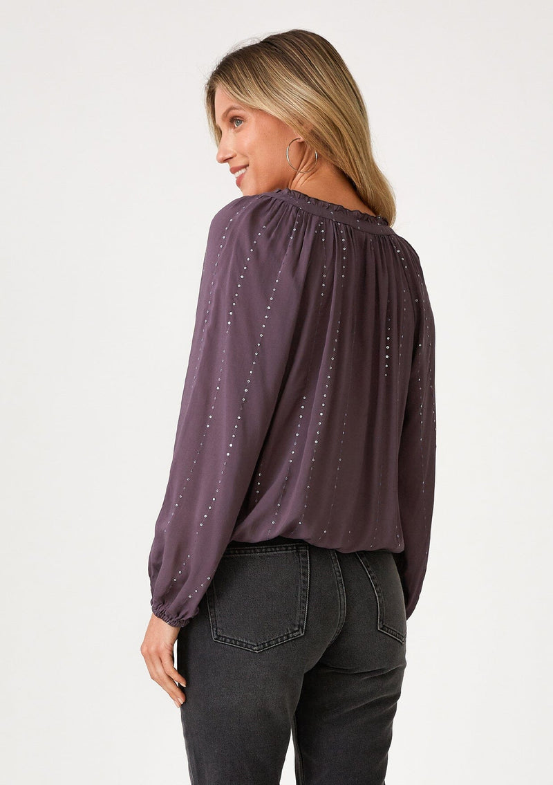 [Color: Dusty Plum] A back facing image of a blonde model wearing a dusty purple bohemian blouse with a sparkly sequined stripe. With long raglan sleeves, a split v neckline with tassel ties, and a ruffled elastic hemline. 